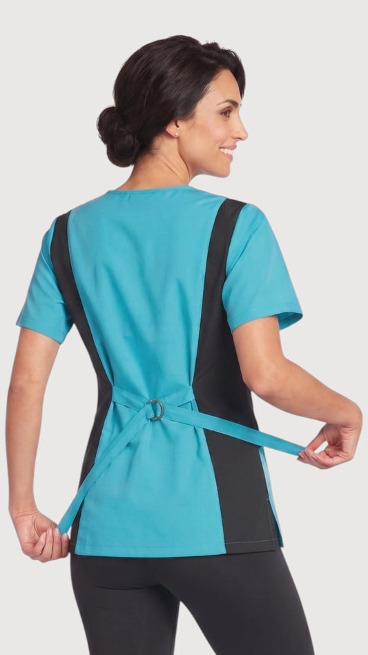 Back view of a brunette woman modeling the Ladybird Line 210 turquoise scrub tunic top, showing the black accent panels and adjusting the turquoise back tie belt, paired with black leggings against a solid light grey studio background.