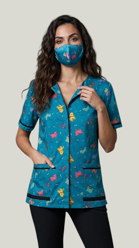 Front view of a woman with long dark wavy hair modeling the Ladybird Line 23 blue dog design pet grooming face mask, paired with a matching short-sleeve grooming tunic top and black pants against a light background.