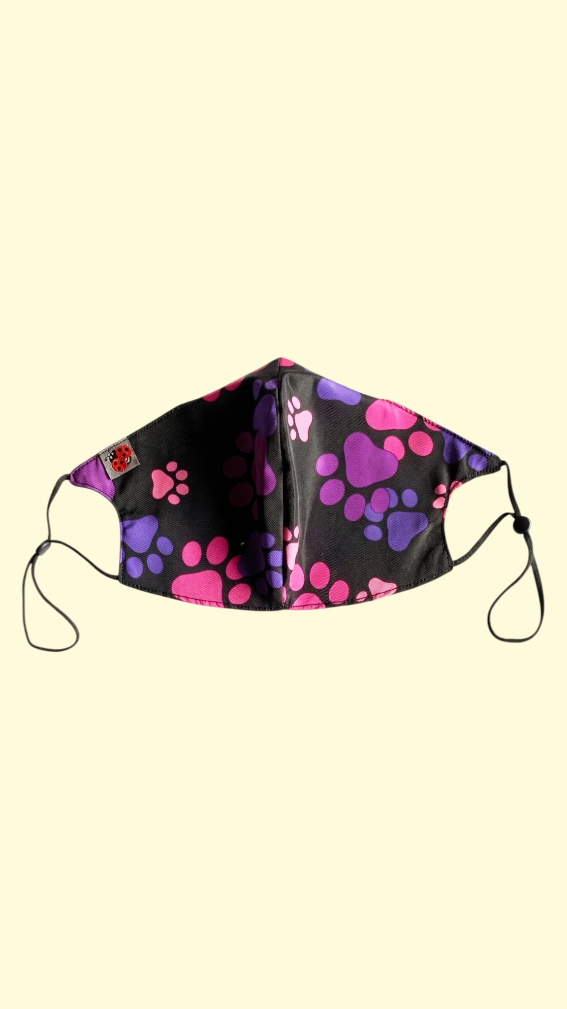 Ladybird Line 27 pet grooming face mask with a vibrant pink and purple paw print pattern on black fabric, featuring adjustable elastic ear loops shown against a solid pale yellow background.
