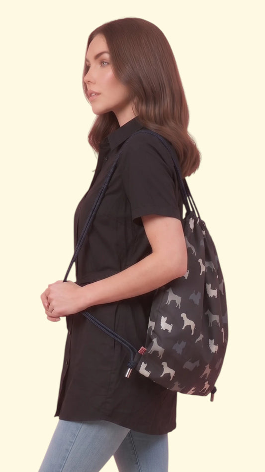 Side profile view of a brunette woman modeling the Ladybird Line 340 drawstring backpack featuring a black, grey, and white Scottish Terrier dog print, paired with a black button-up short-sleeve tunic and blue jeans against a solid off-white background.