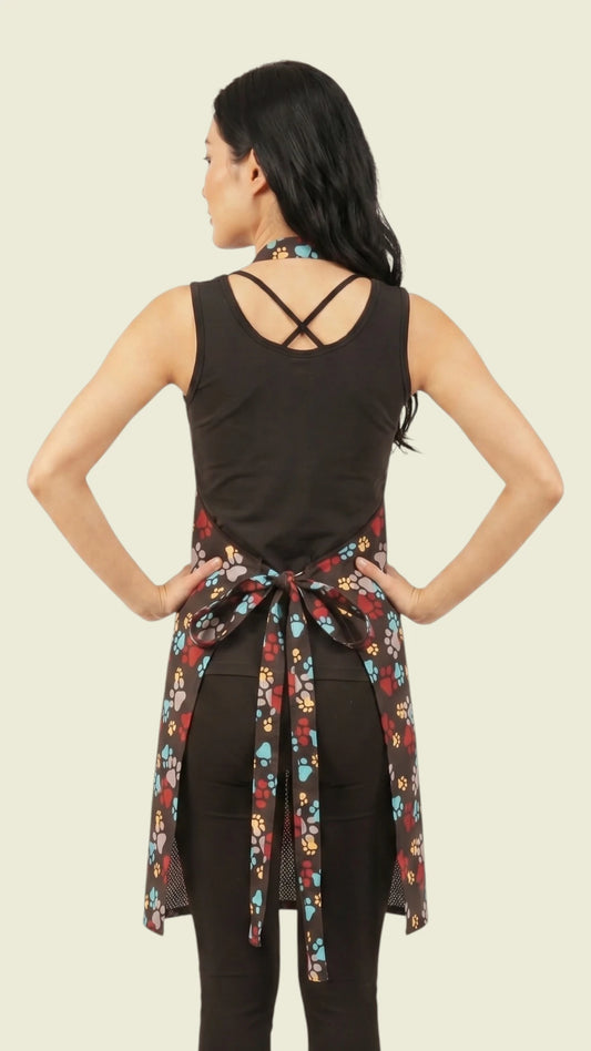 Rear view of a woman with long dark hair posing with hands on hips, modeling the Ladybird Line 601 waterproof pet grooming apron in a multicolor paw print pattern, showing the tie-waist bow and breathable black mesh backing over a black sleeveless top and dark pants against a light cream background.