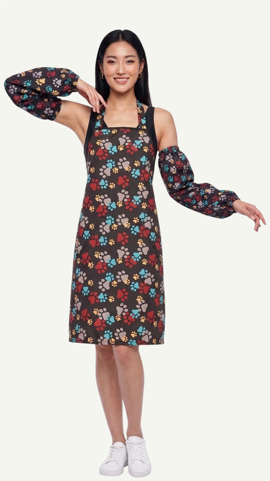 Full-length front view of a smiling woman modeling the Ladybird Line 603 waterproof paw print dog bathing sleeves, paired with a matching paw print pet grooming apron dress against an off-white background.
