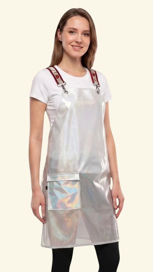 Three-quarter side view of a young woman smiling while modeling the Ladybird Line 111 Waterproof Clear Sparkle Crossover Apron, featuring a clear sparkle plastic apron with red signature crossover straps and front flap pockets, worn over a white t-shirt and black pants against a light beige background.