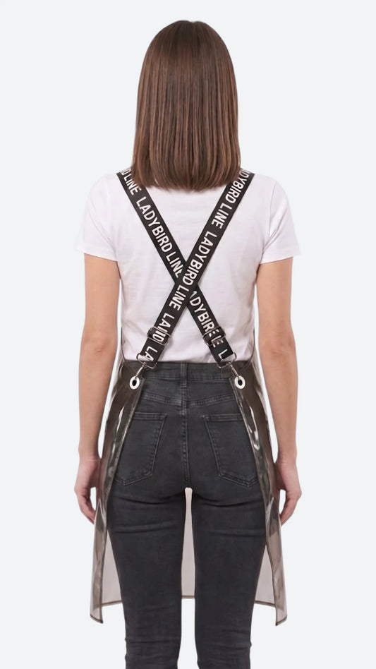 Back view of a dark-haired woman modeling the Ladybird Line 113 Waterproof Black Clear Crossover Apron, showing the crisscross black signature straps printed with white 'LADYBIRD LINE' text over a white t-shirt and dark pants against a solid white background.