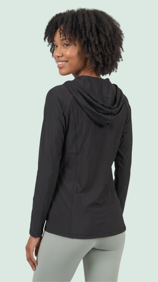 Back three-quarter view of a woman with short curly dark hair modeling the Ladybird Line 122 Black Shadow Hooded Hair & Bleach Resistant Grooming Jacket with the hood down, worn over light gray leggings against a light green background.