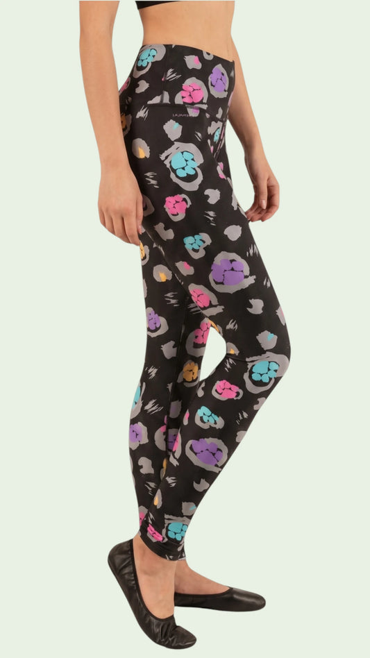 Side profile view of a woman modeling the Ladybird Line 126 Paw Print Shadow Hair & Bleach Resistant Grooming Leggings featuring a colorful pink, teal, purple, and yellow paw print pattern, worn with black flats against a light green background.