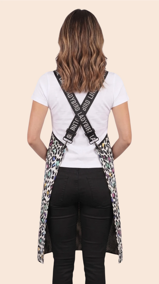 Back view of a dark-haired woman modeling the Ladybird Line 137 Multi Color Leopard Print Waterproof Signature Strap Crossover Apron, showing the crisscross black signature straps printed with white 'LADYBIRD LINE' text over a white t-shirt and black pants against a solid pale peach background.