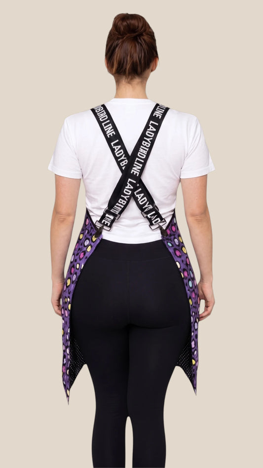 Back view of a woman modeling the Ladybird Line 139 Lilac Mini Paw Print Waterproof Signature Strap Crossover Apron, displaying the crisscross back straps with Ladybird Line branding, worn over a white t-shirt and black pants against a light beige background.