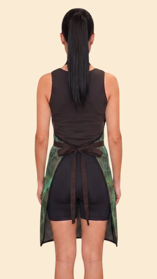 Back view of a woman with a dark ponytail modeling the Ladybird Line 142 Green Sparkle Waterproof & Bleach Resistant Apron, tied neatly at the waist over a black top and shorts against a pale beige background.