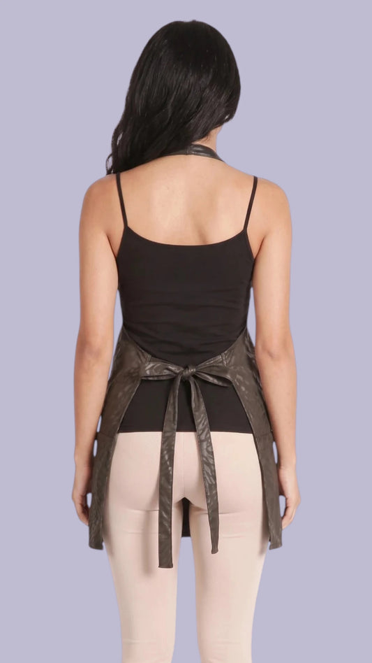 Back view of a dark-haired woman modeling the Ladybird Line 145 Black Leopard Print Water & Bleach Resistant Grooming Apron, tied neatly at the waist over a black camisole and cream leggings against a soft lavender background.