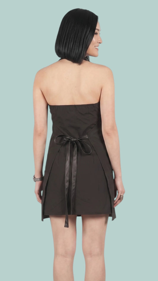 Back view of a woman with dark hair modeling the Ladybird Line 150 Rhinestone Zipper Pro Water Resistant Salon Grooming Apron in black, tied neatly at the waist with dark straps against a solid light blue background.