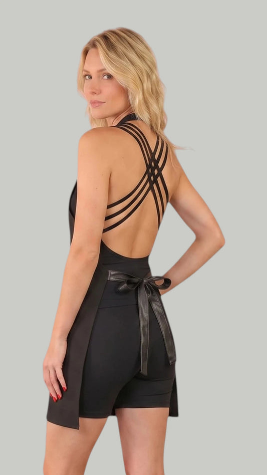 Back three-quarter view of a blonde woman modeling the Ladybird Line 270 Rhinestone Zipper Water Resistant Styling Apron, showing the crisscross back strap design, an open lower back, and a shiny faux-leather waist tie belt tied in a bow, set against a solid light grey background.
