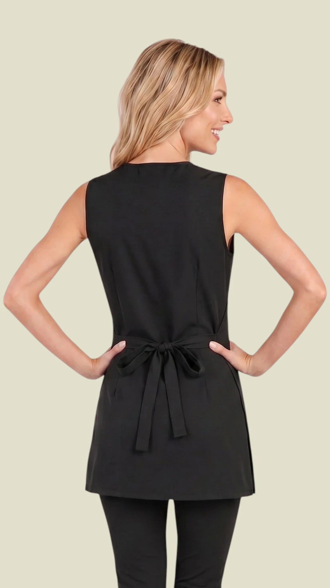 Back view of a blonde woman posing in the Ladybird Line 345 Metal Stud Water Resistant Cobbler Apron, showing the solid black sleeveless cobbler apron back with a side tie belt secured in a bow at the waist, worn over black leggings against a solid light beige background.