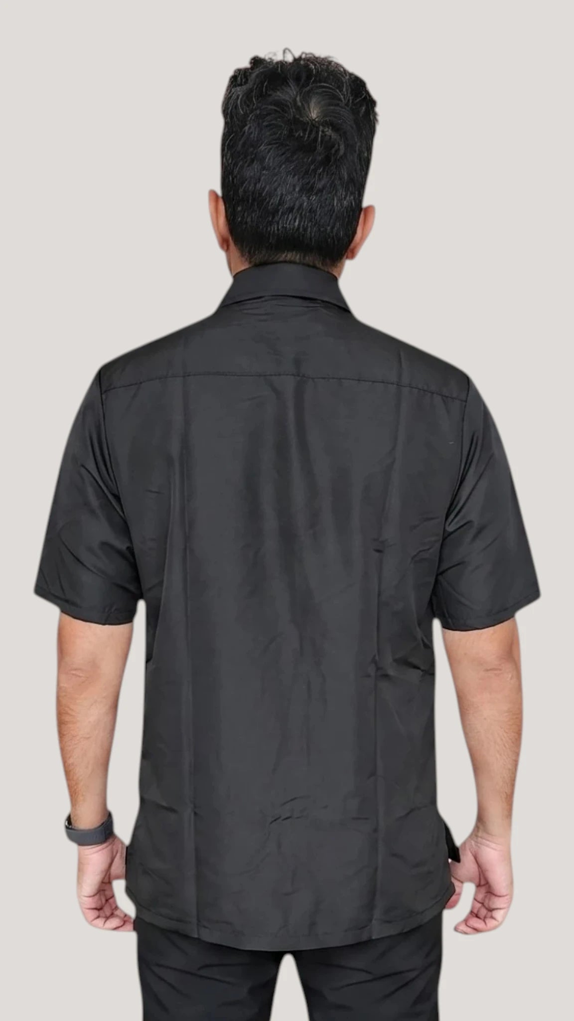 Back view of a dark-haired man modeling the Ladybird Line 420 Men's Terrier Print Water Resistant Grooming Jacket, showing the solid black short-sleeve jacket back with a crisp collar, worn over black pants against a solid light grey background.