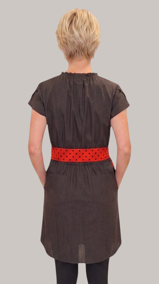 Back view of a blonde woman modeling the Ladybird Line 444 Classic Black Water Resistant Scarf Belt Professional Dress, showing the back elastic waist and red polka dot scarf belt worn over dark pants against a solid light grey background.