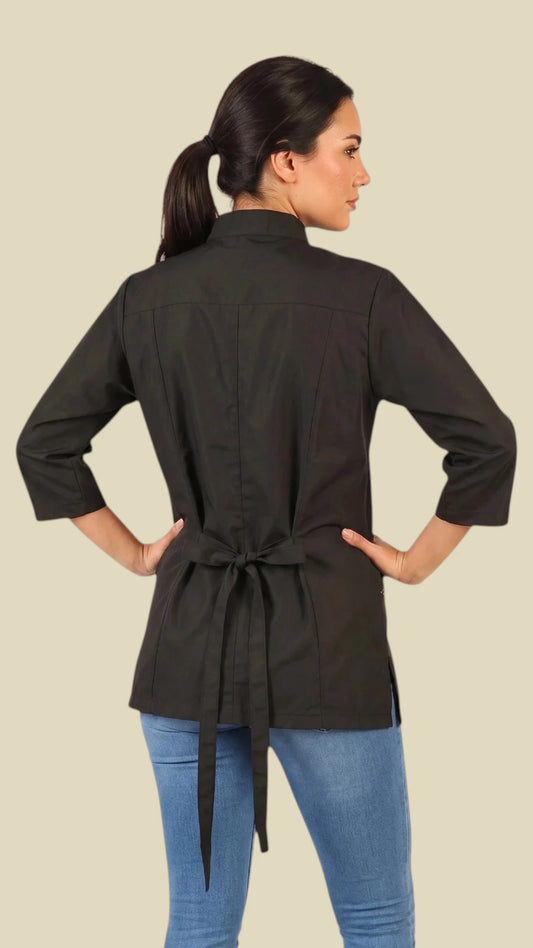 Back view of a dark-haired woman looking back over her shoulder while modeling the Ladybird Line 512 Women's Black 3/4 Sleeve Water Resistant Stylist Jacket, showing the black jacket back with 3/4 sleeves, mandarin collar, and adjustable tie belt tied into a bow at the back waist, worn over blue jeans against a solid light beige background.