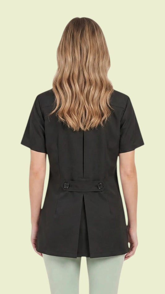 Back view of a blonde woman wearing light sage green pants while modeling the Ladybird Line 530 Pro Salon Water Resistant & Bleach Resistant Styling Jacket, showing the black short-sleeve jacket's pleated back design and decorative back belt with buttons against a solid pale yellow background.