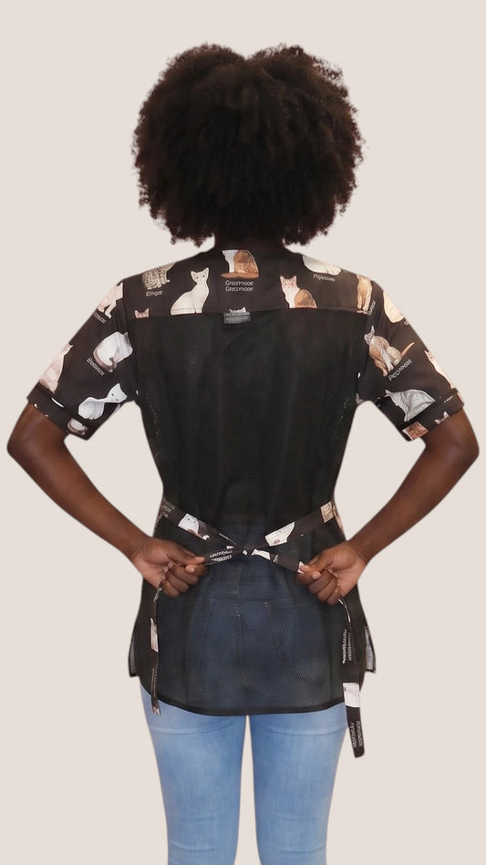 Back view of a dark-skinned woman with curly hair modeling the Ladybird Line 545 The Catty Groomer Waterproof & Bleach Resistant Grooming Jacket, showing the black mesh back panel, adjustable waist tie belt, and printed shoulder panels, worn with blue jeans against a solid off-white background.