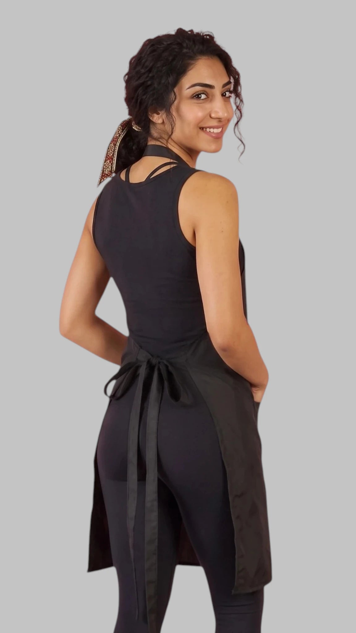 Front view of a dark-haired woman smiling with hands in pockets modeling the Ladybird Line 600 Women's Classic Black Waterproof & Hair Resistant Bather's Grooming Apron, featuring a solid black apron with an adjustable neck strap and deep side pockets, worn over a black tank top and black leggings against a light grey background.