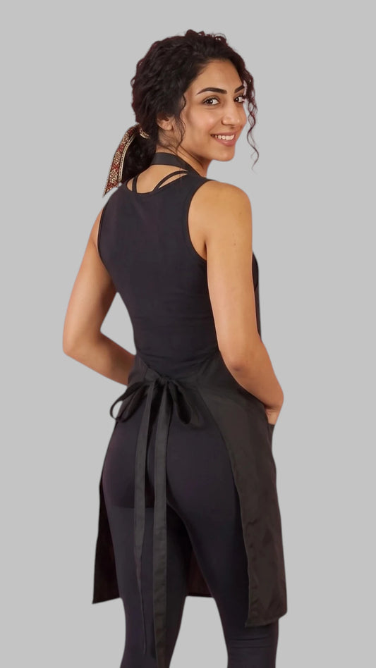Front view of a dark-haired woman smiling with hands in pockets modeling the Ladybird Line 600 Women's Classic Black Waterproof & Hair Resistant Bather's Grooming Apron, featuring a solid black apron with an adjustable neck strap and deep side pockets, worn over a black tank top and black leggings against a light grey background.