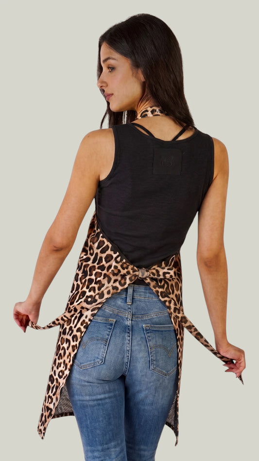 Back view of a woman with dark hair modeling the Ladybird Line 602 Leopard Print Waterproof & Bleach Resistant Apron, showing the back tie closure, inner mesh lining, and side towel loop, worn with a black tank top and blue jeans against a light grey background.