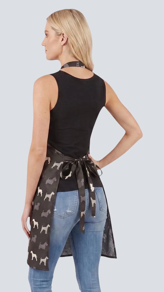 Front view of a smiling blonde woman posing with hand on hip modeling the Ladybird Line 613 Terrier Breed Print Waterproof & Bleach Resistant Grooming Apron, featuring a full-coverage black apron with a repeating white and grey terrier silhouette print, adjustable neck band, and deep side pockets, worn over blue jeans against a light grey background.