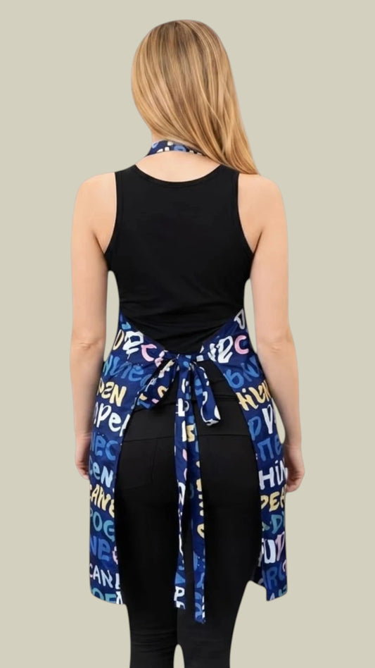 Back view of a blonde woman wearing a black tank top and black leggings while modeling the Ladybird Line 620 Unisex Universal Dog Print Waterproof & Bleach Resistant Grooming Apron, showing the navy blue back tie belt with multilingual "dog" typography print tied around the waist against a light beige background.