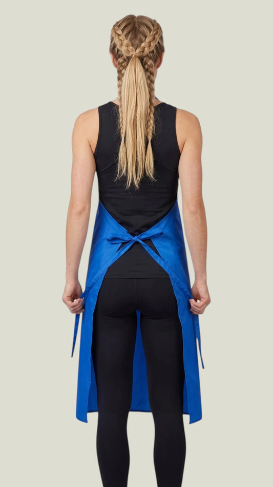 Back view of a blonde woman with braided hair modeling the Ladybird Line 680 Bathers Long Royal Blue Waterproof Bathing Apron, showing the crisscross back tie belt cinched at the waist, worn over a black sleeveless top and black leggings against a light beige background.
