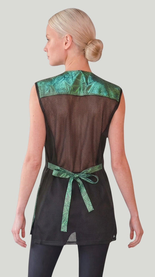 Front view of a blonde woman posing with hands in pockets modeling the Ladybird Line 700 Sparkling Green Waterproof & Bleach Resistant Mesh Back Grooming Vest, featuring a sleeveless grooming vest with a sparkling green color block geometric pattern, covered front zipper, black side panels, and deep pockets, worn over black pants against a light grey background.