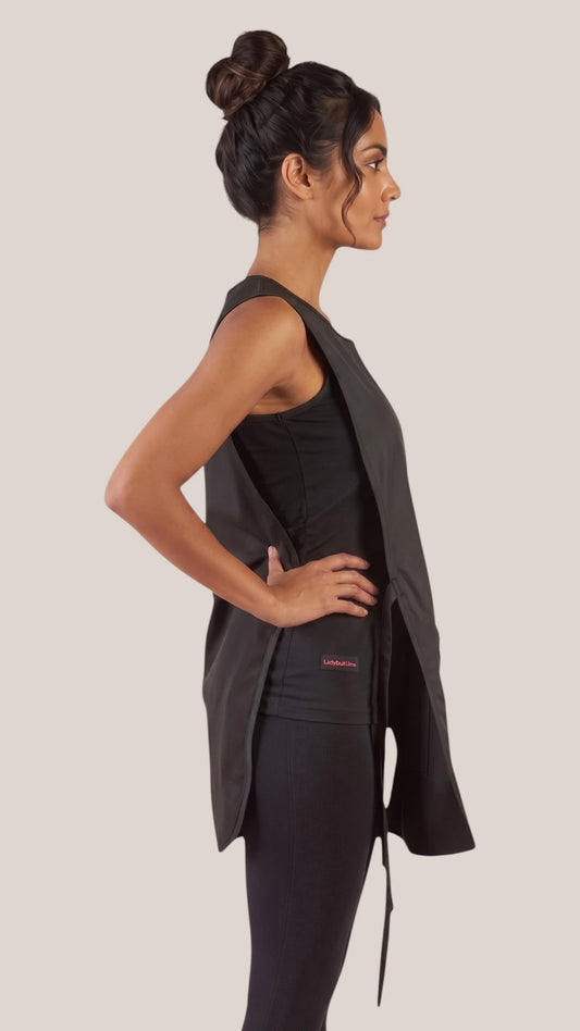 Side profile view of a dark-haired woman in a hair bun modeling the Ladybird Line 775 Waterproof Hair Resistant & Bleach Resistant Regular Size Cobbler Apron, showing the open side profile with hanging tie belts, worn over a black top and black leggings against a light beige background.