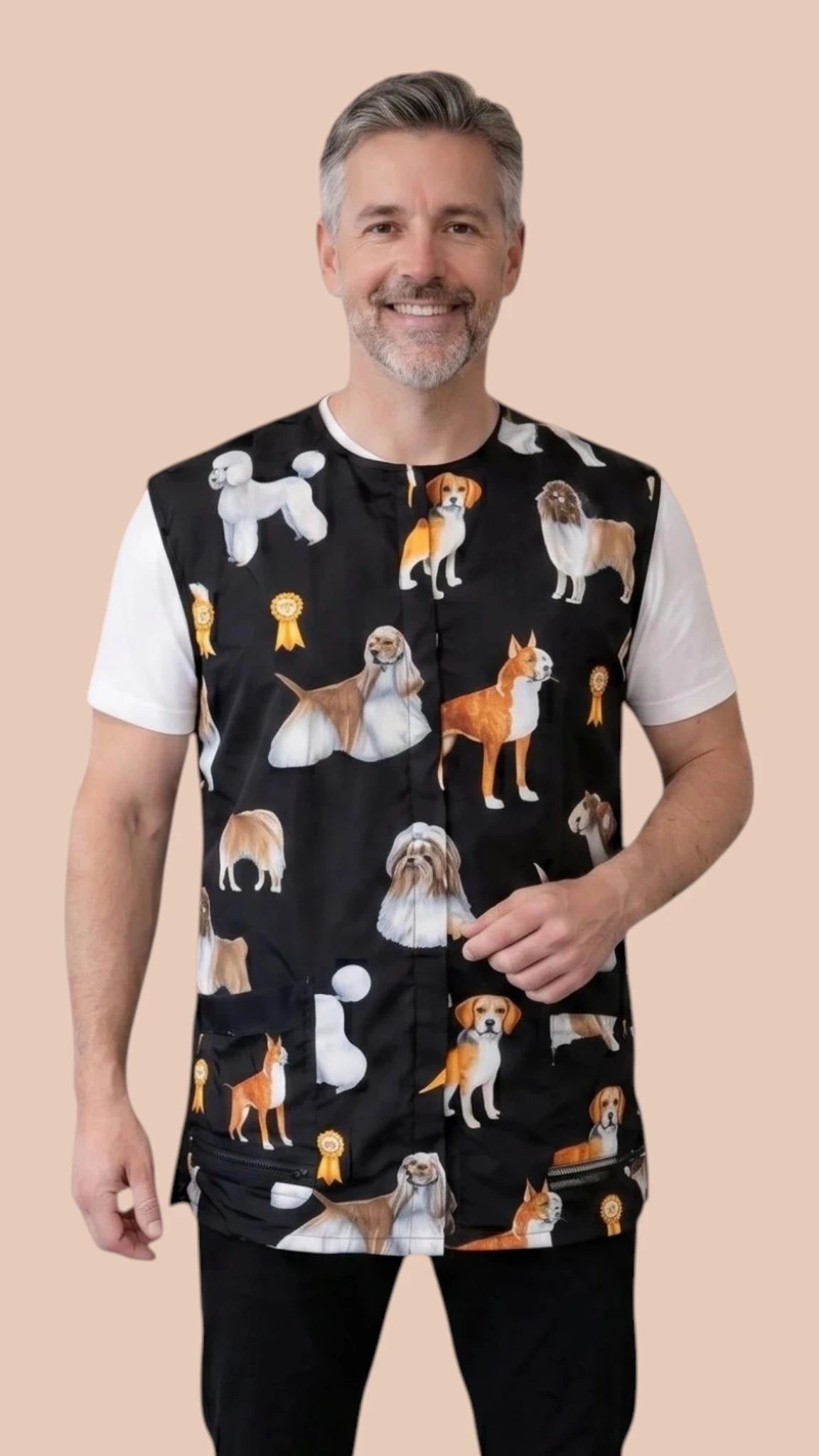 Front view of a smiling man with short grey hair modeling the Ladybird Line 777 Dog Groups Waterproof & Bleach Resistant Grooming Vest with Mesh Back, featuring a sleeveless black grooming vest with an all-over print of various dog breed groups, worn over a white t-shirt and black pants against a solid light beige background.