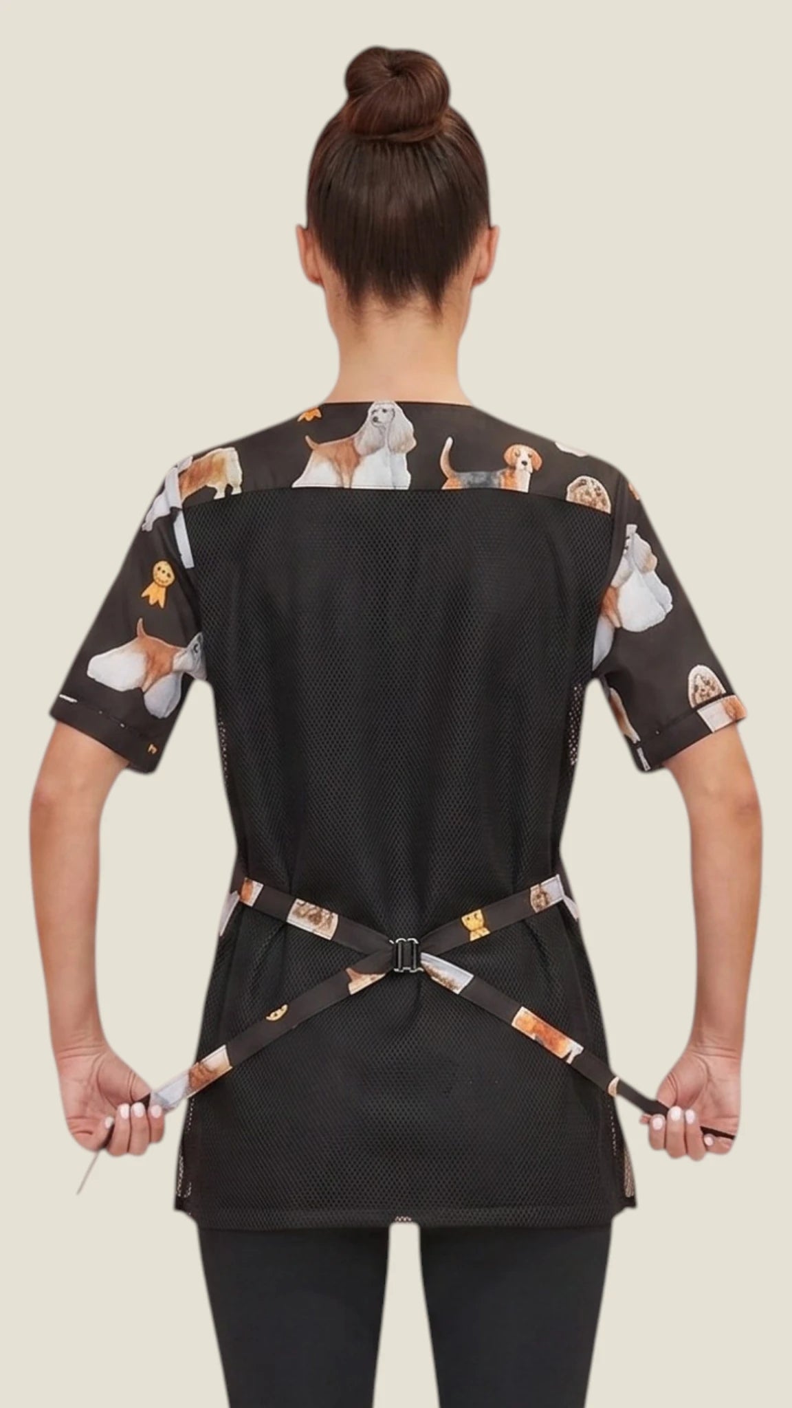 Back view of a dark-haired woman in a hair bun modeling the Ladybird Line 803 Dog Groups Water Resistant & Bleach Resistant Stylist Jacket with Mesh Back, showing the black mesh back panel, printed upper shoulder yoke, short sleeves, and printed back tie belt, worn over dark pants against a light beige background.