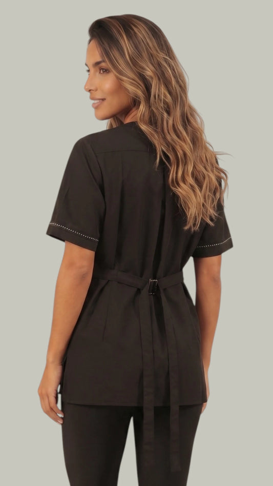 Back view of a brown-haired woman modeling the Ladybird Line 850 Rhinestone Trim Water, Bleach & Hair Resistant Styling Jacket, showing the back tie belt cinched at the waist and contrast stitching accents, worn with black pants against a solid light grey background.