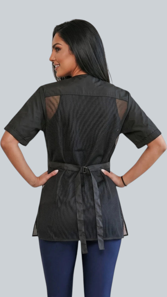 Front view of a dark-haired woman posing with hands in pockets modeling the Ladybird Line 950 Classic Black Waterproof & Bleach Resistant Grooming Smock, featuring a short-sleeve black grooming jacket with covered front zipper and front pockets, worn over dark blue pants against a light grey background.