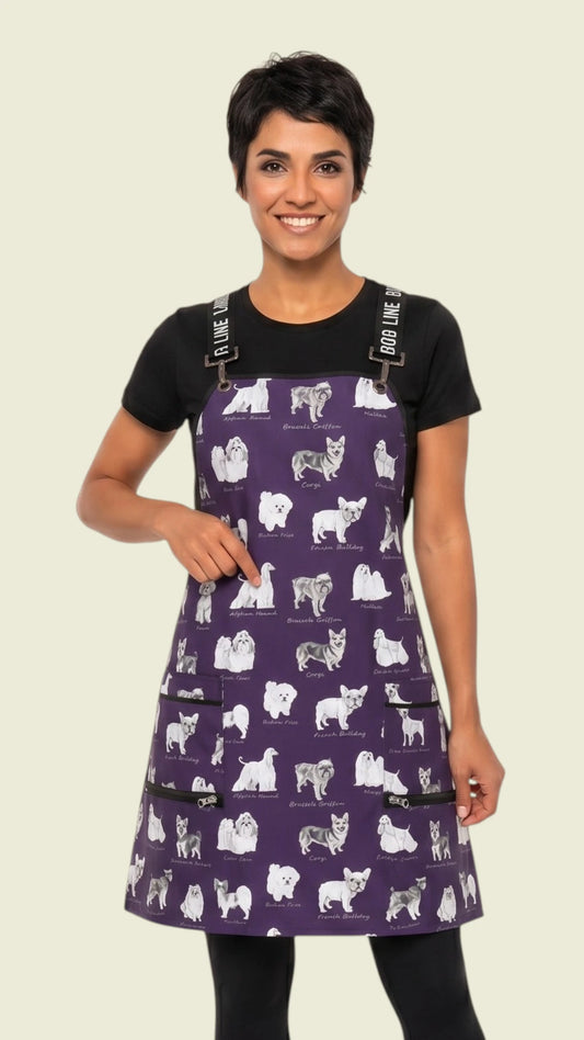 Three-quarter length front view of a smiling woman with short dark hair modeling the Ladybird Line 134 Puppy Love sketch waterproof pet grooming apron in deep purple with puppy breed print, worn over a black t-shirt and dark pants against a light background.