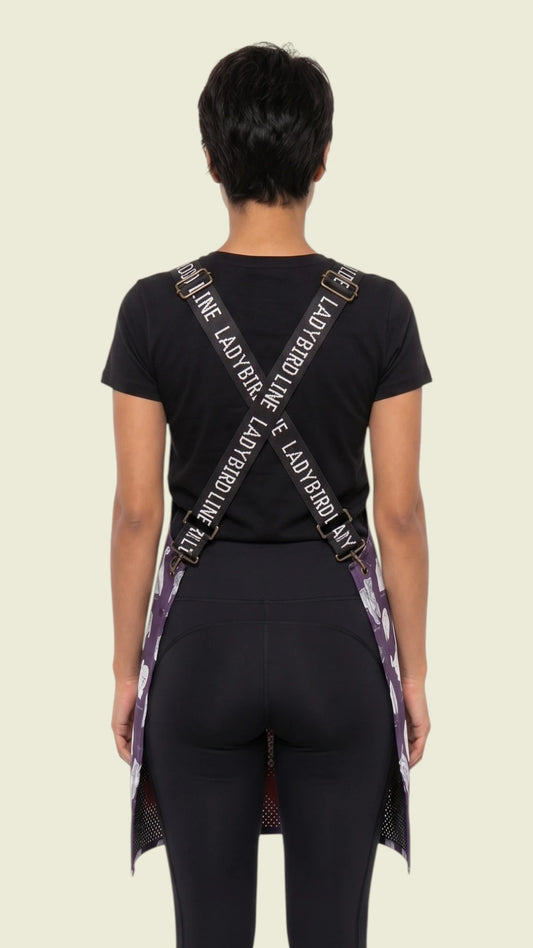 Rear view of a woman with short brunette hair modeling the Ladybird Line 134 Puppy Love sketch waterproof pet grooming apron, showing the crisscross back straps with Ladybird Line branding and deep purple puppy print over a black t-shirt and black leggings against a light background.