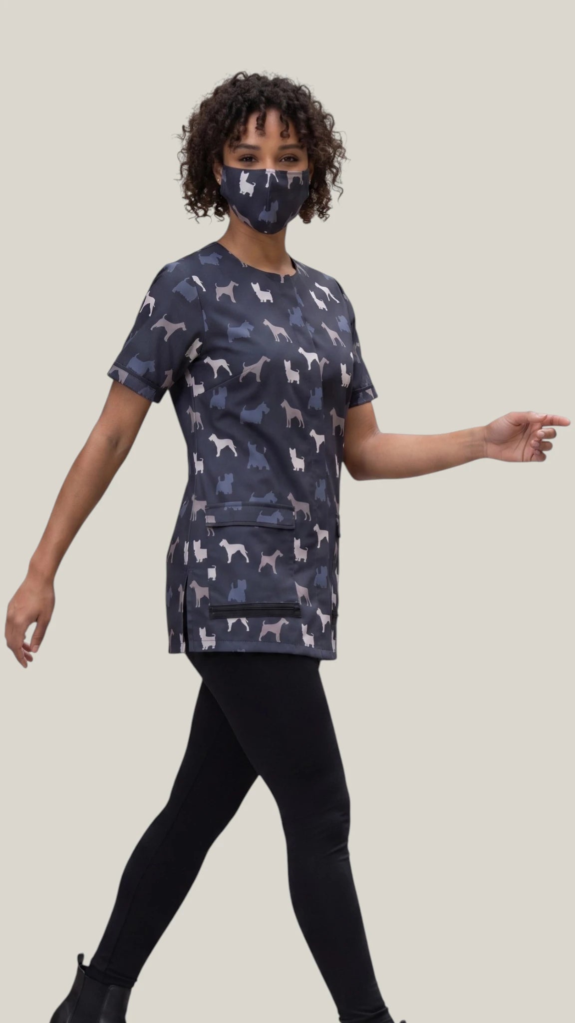 Full-length side-angle view of a woman with short curly dark hair modeling the Ladybird Line 14 terrier breed print pet grooming face mask, paired with a matching short-sleeve grooming tunic top and black leggings against a solid light gray background.