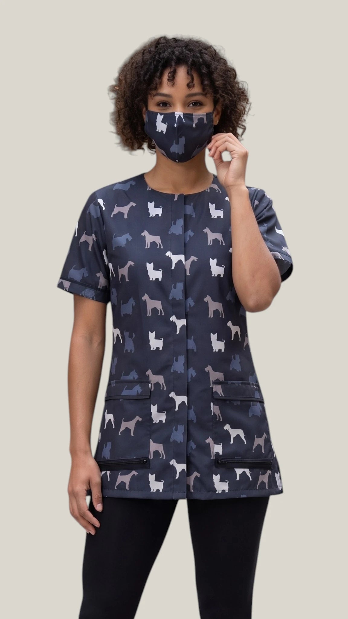 Front view of a woman with short curly dark hair modeling the Ladybird Line 14 terrier breed print pet grooming face mask, paired with a matching short-sleeve grooming tunic top and black pants against a solid light background.