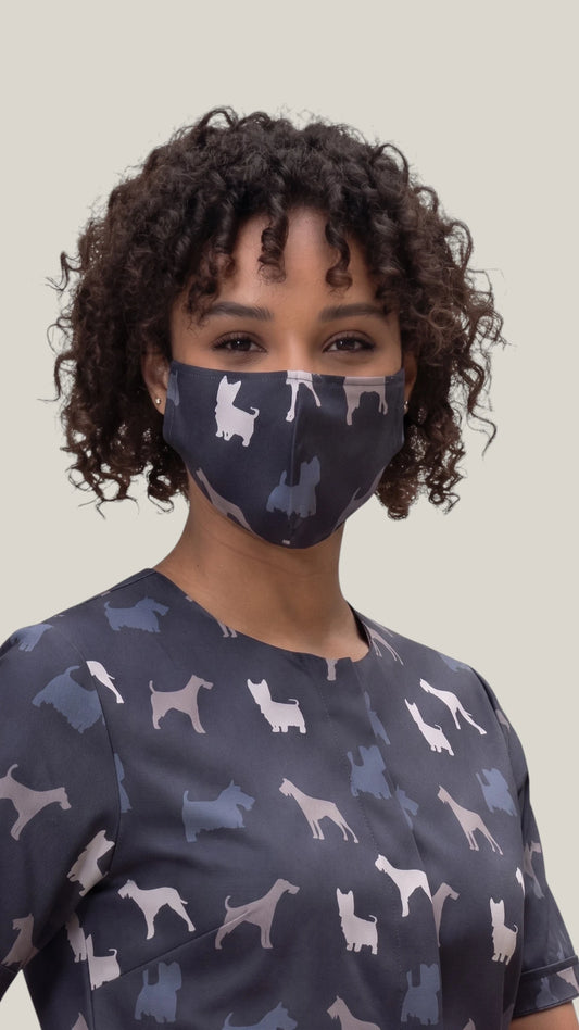 Close-up front view of a woman with short curly dark hair modeling the Ladybird Line 14 navy blue pet grooming face mask with a terrier breed silhouette pattern, paired with a matching grooming top against a light gray background.