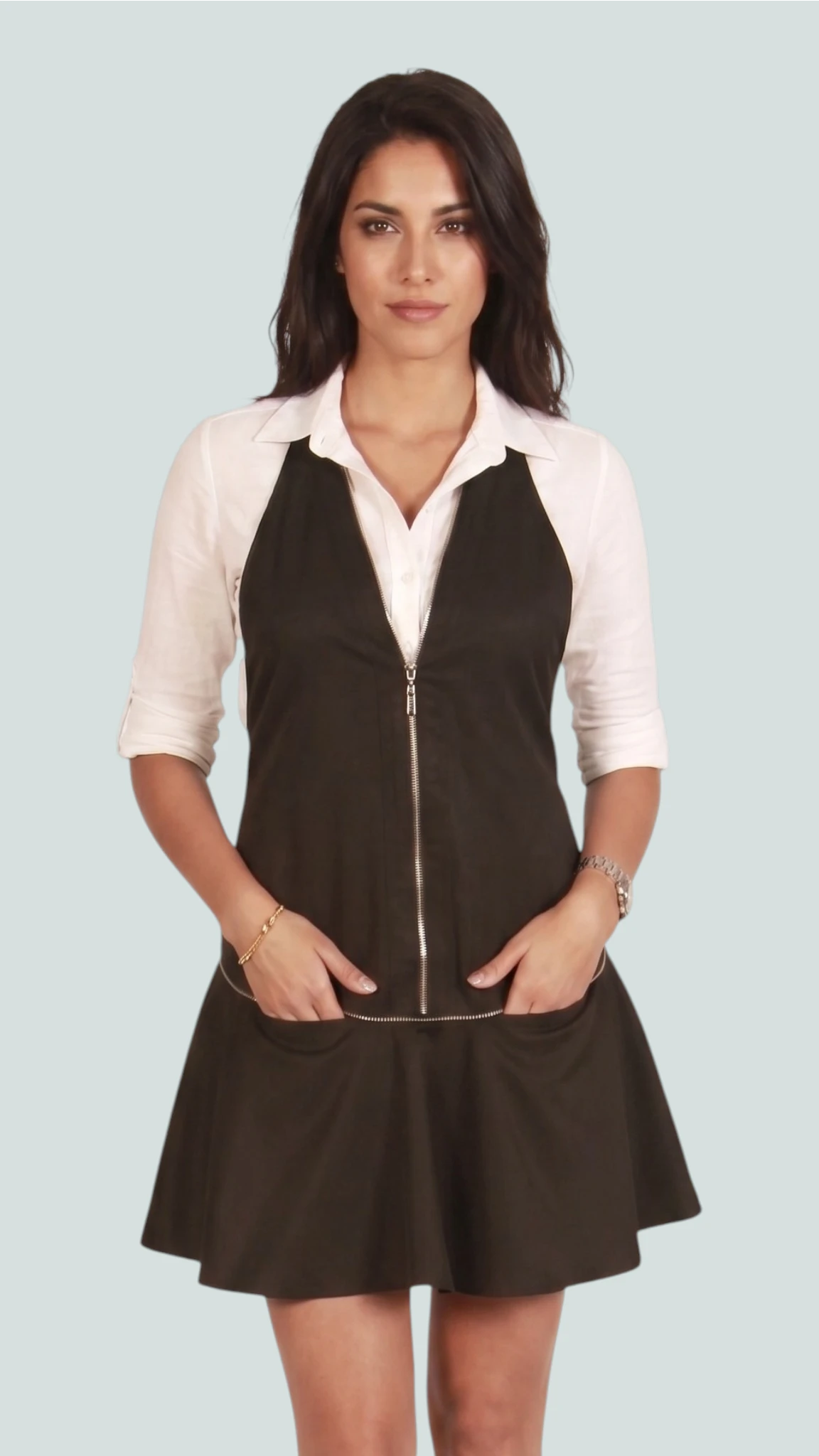 Front view of a brunette woman modeling the Ladybird Line 140BK black vintage dress-style stylist apron with a front zipper and flared pleated hem, hands in front pockets, worn over a white collared blouse against a light studio background.
