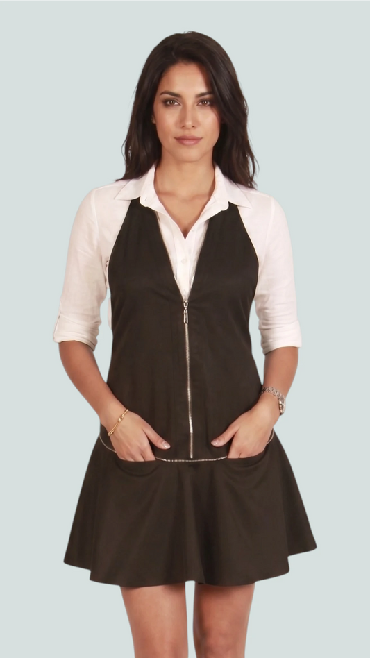 Front view of a brunette woman modeling the Ladybird Line 140BK black vintage dress-style stylist apron with a front zipper and flared pleated hem, hands in front pockets, worn over a white collared blouse against a light studio background.