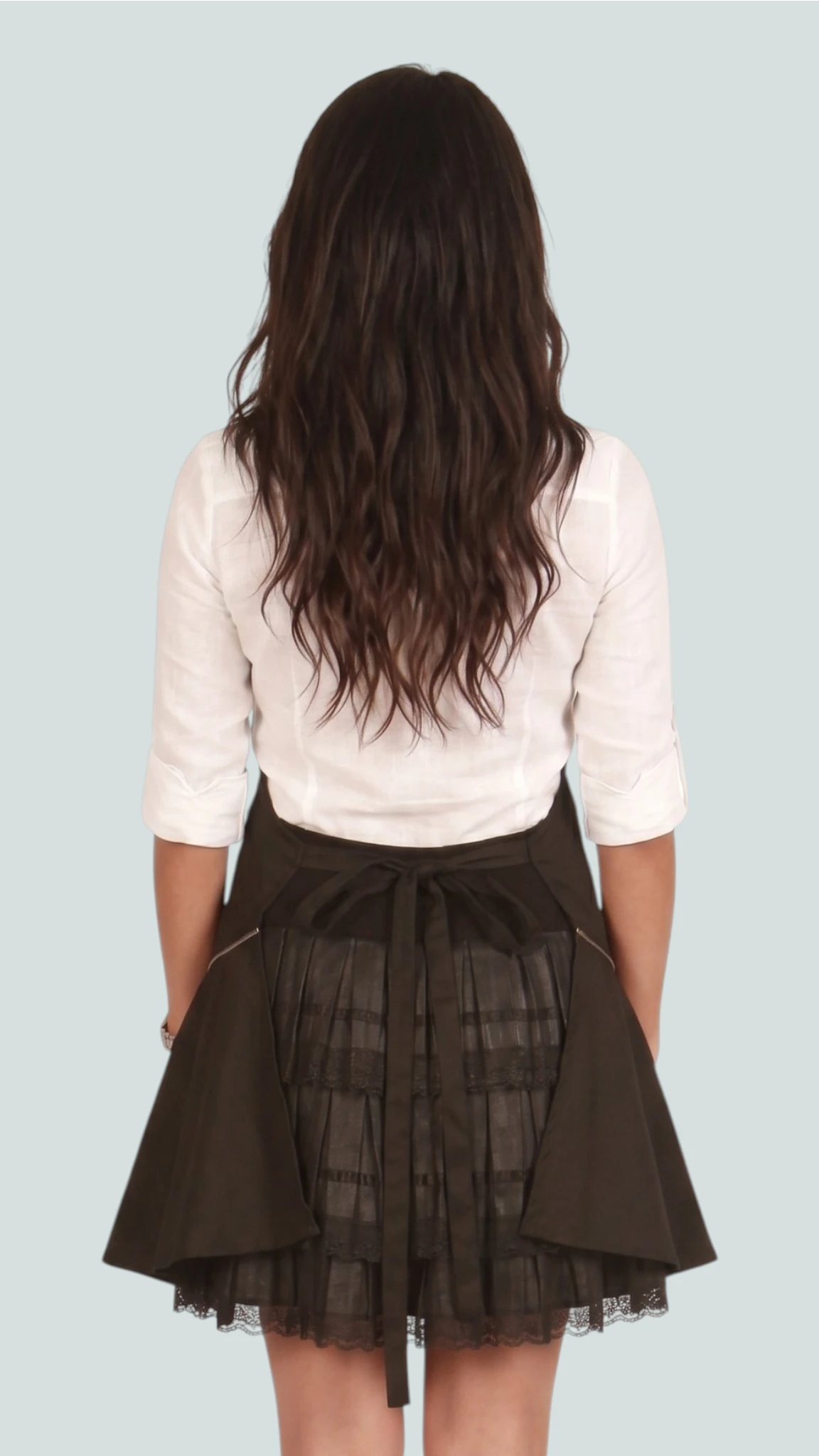 Back view of a woman with long dark wavy hair modeling the Ladybird Line 140BK black vintage dress stylist apron, highlighting the tied waist bow and pleated flared lower skirt over a white top against a solid light blue background.