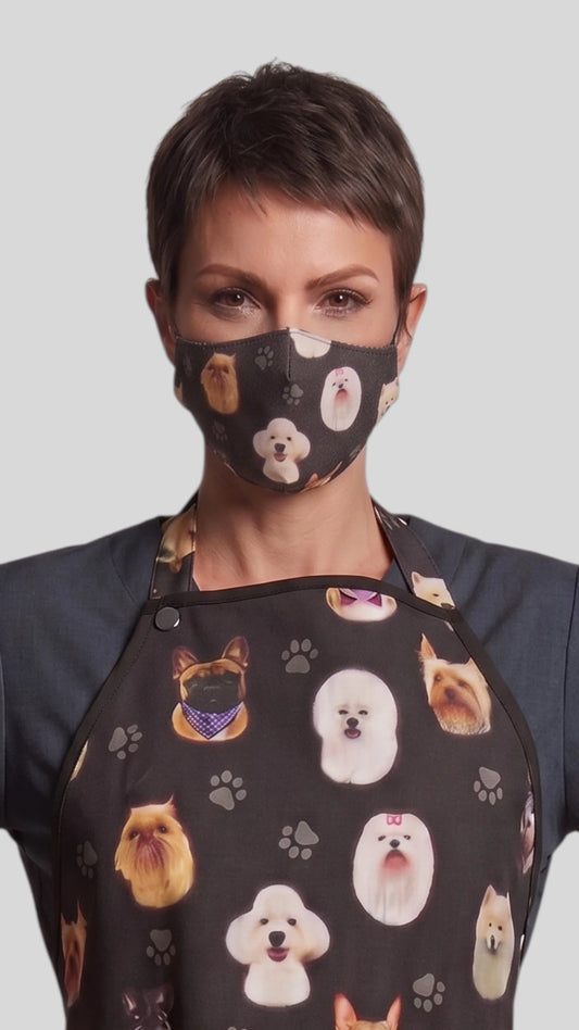 Close-up front view of a woman with short dark hair modeling the Ladybird Line 21 black pet groomer face mask featuring dog breed head illustrations, paired with a matching dog print grooming apron against a light gray background.