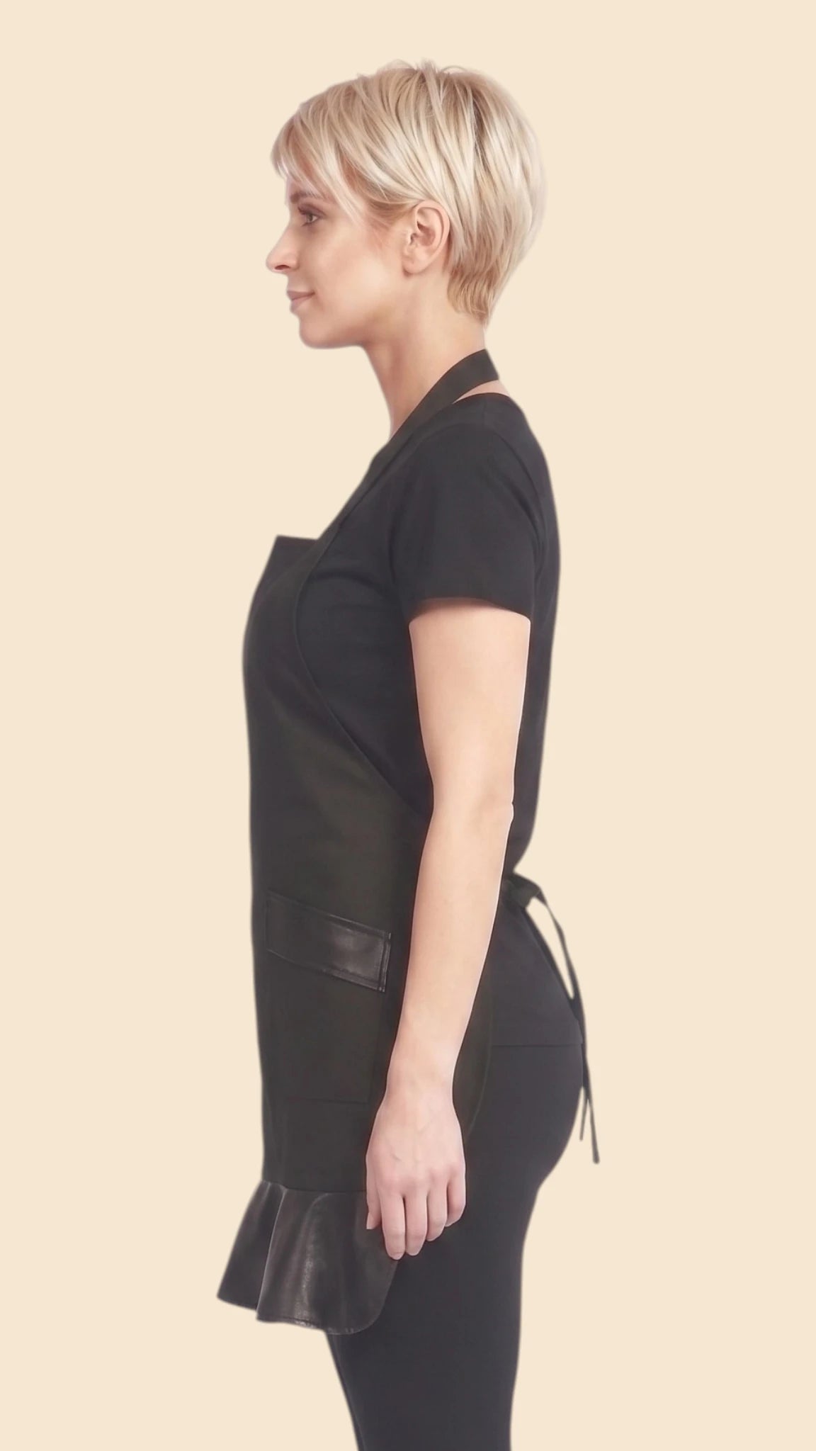 Side profile view of a blonde woman with short hair modeling the Ladybird Line 215 black stylist cocktail apron, showing the faux leather pleated hem and side silhouette, worn over a black t-shirt and dark pants against an off-white studio background.