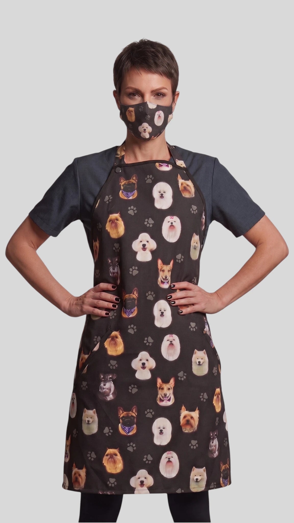 Three-quarter length front view of a woman with short dark hair posing with hands on hips, modeling the Ladybird Line 21 dog breed print pet groomer face mask paired with a matching dog breed print grooming apron over a short-sleeve top against a gray background.
