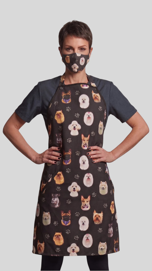 Three-quarter length front view of a woman with short dark hair posing with hands on hips, modeling the Ladybird Line 21 dog breed print pet groomer face mask paired with a matching dog breed print grooming apron over a short-sleeve top against a gray background.