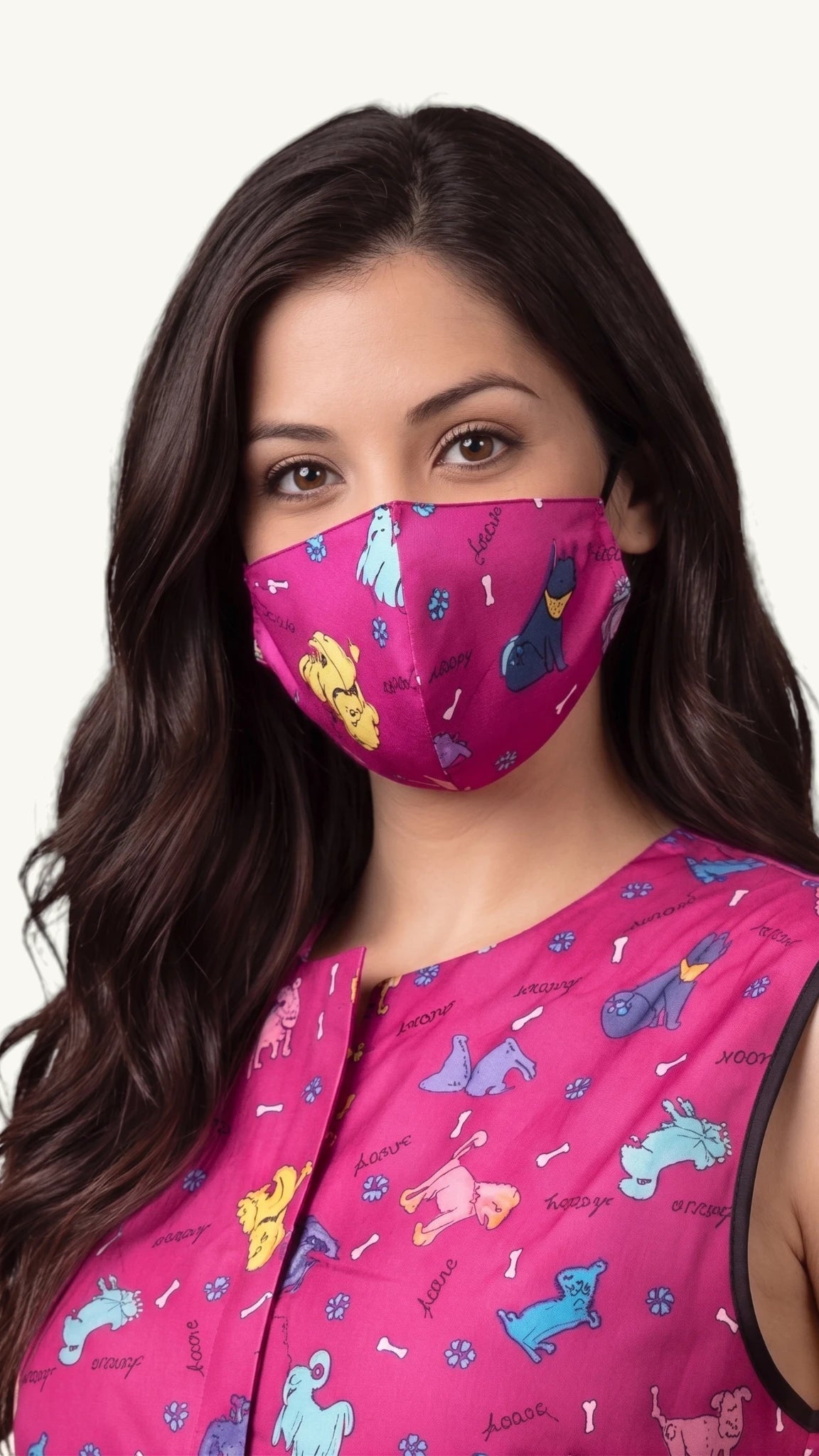Close-up front view of a woman with long dark wavy hair modeling the Ladybird Line 22 pet grooming face mask in a vibrant pink dog print pattern, paired with a matching sleeveless grooming top against a light gray background.