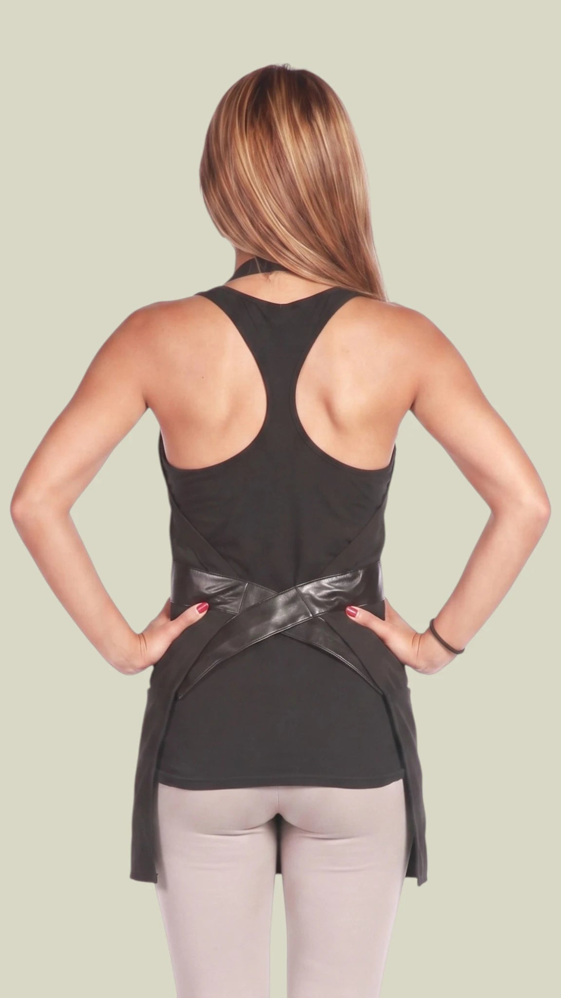 Rear view of a woman with highlighted brown hair posing with hands on hips, modeling the Ladybird Line 223 black V-neck salon styling apron, highlighting the crossover faux leather back belt closure worn over a black racerback tank top and light leggings against a light beige background.