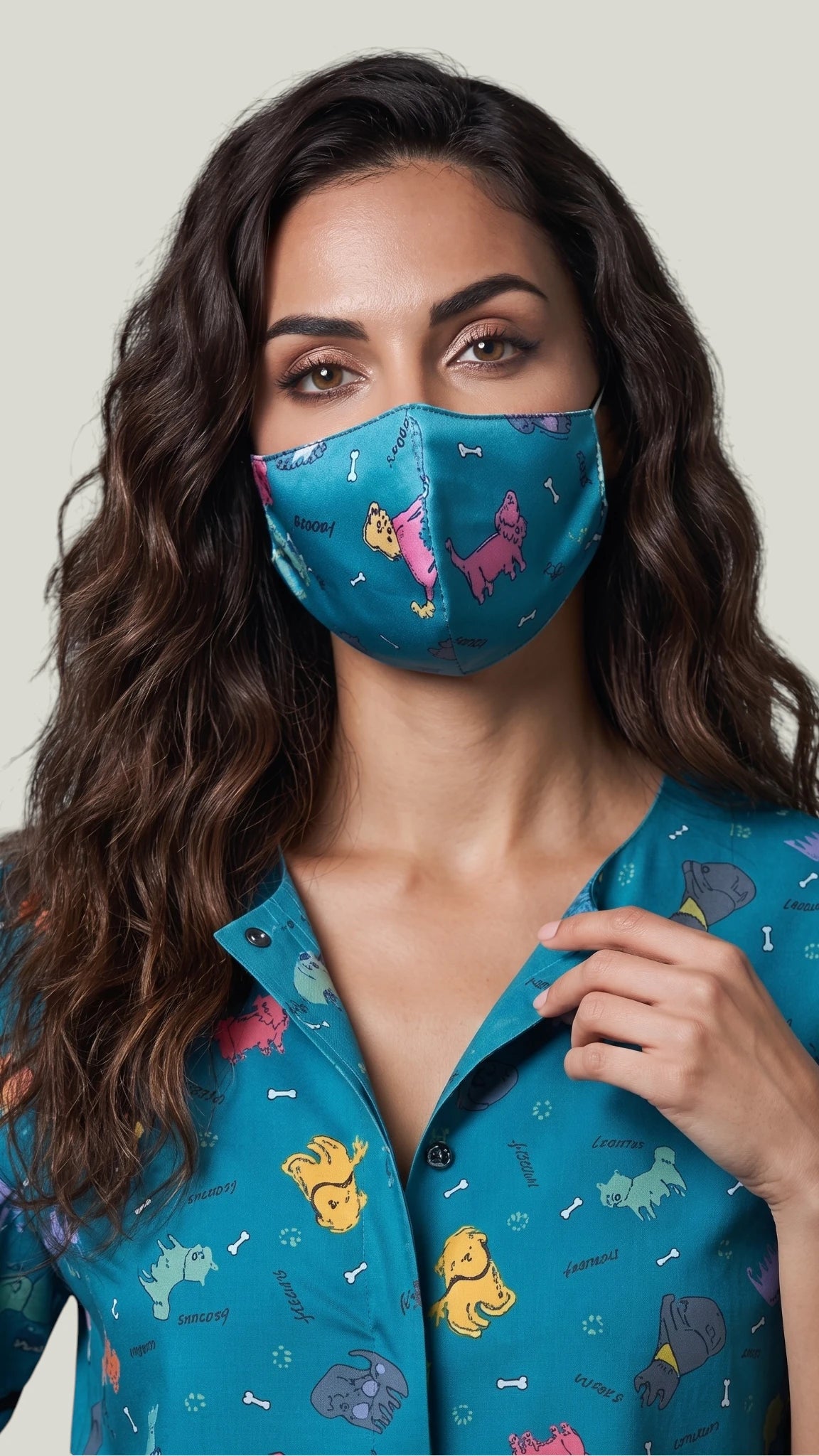 Close-up front view of a woman with long wavy brown hair modeling the Ladybird Line 23 teal blue dog print pet grooming face mask, paired with a matching button-up dog print grooming smock top against a light gray background.