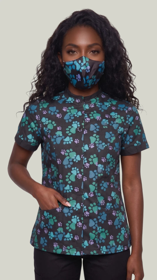 Front view of a woman with long wavy dark hair modeling the Ladybird Line 24 blue paw print pet grooming face mask, paired with a matching short-sleeve grooming top and black pants against a solid light background.